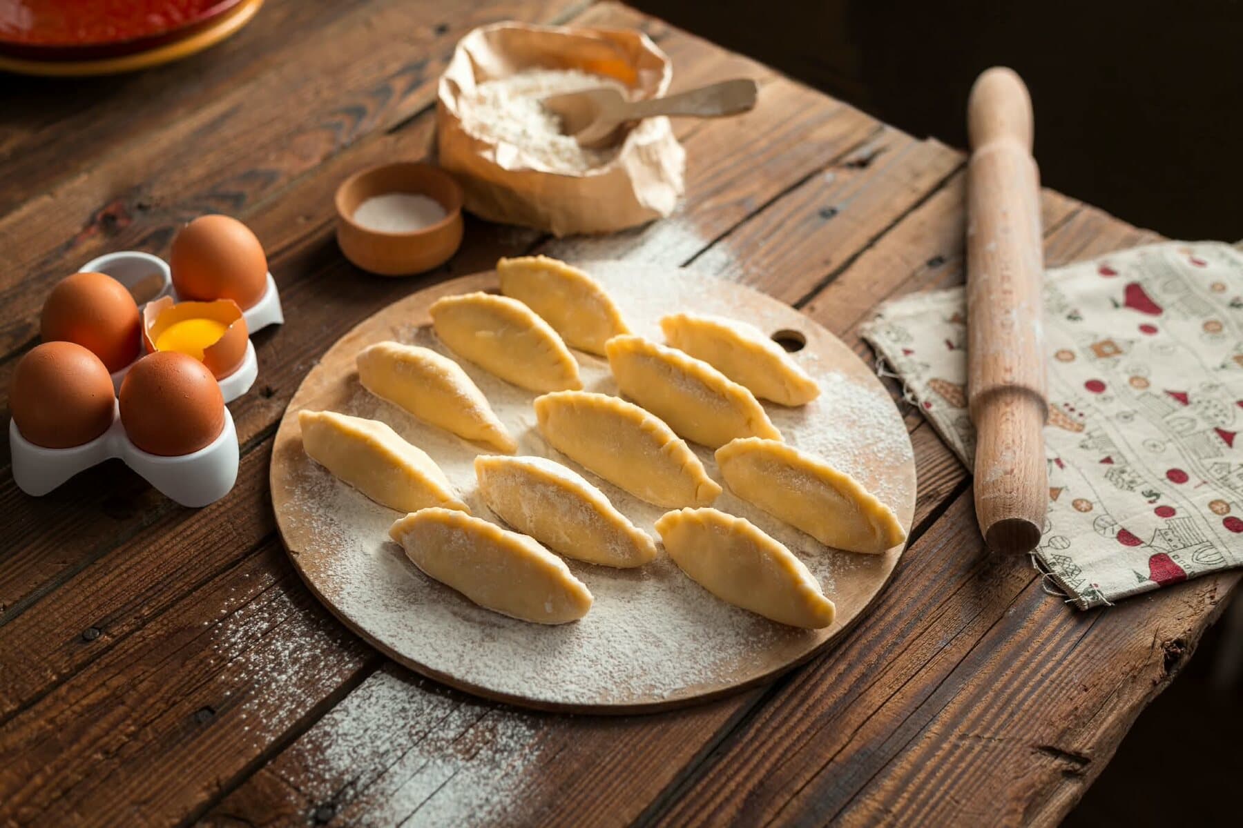 Des ravioli en demi-lune sur une planche farinée, des œufs et le rouleau à côté