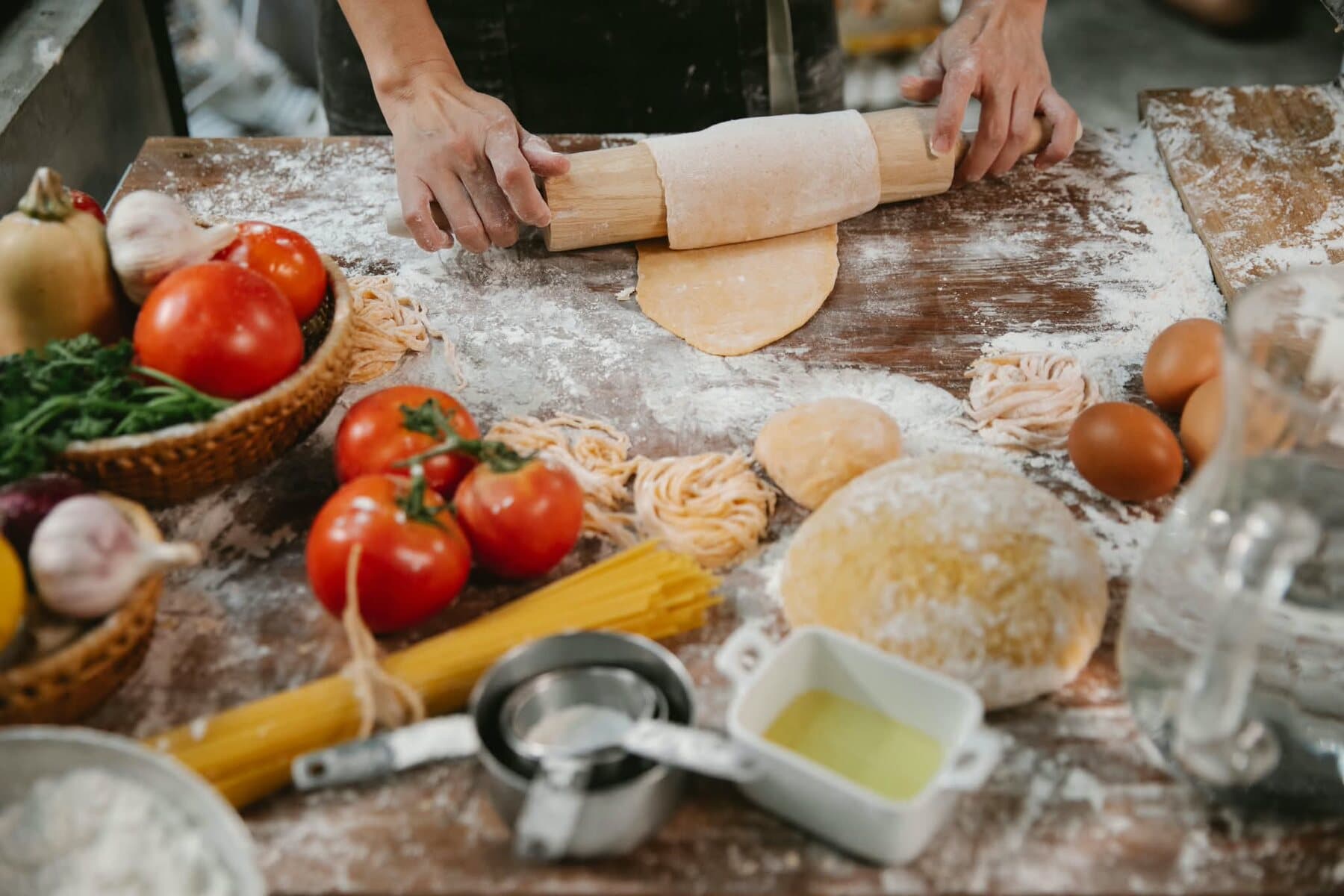 Des mains abaissent la pasta au rouleau, entre les tomates, les nids de tagliatelle et les œufs